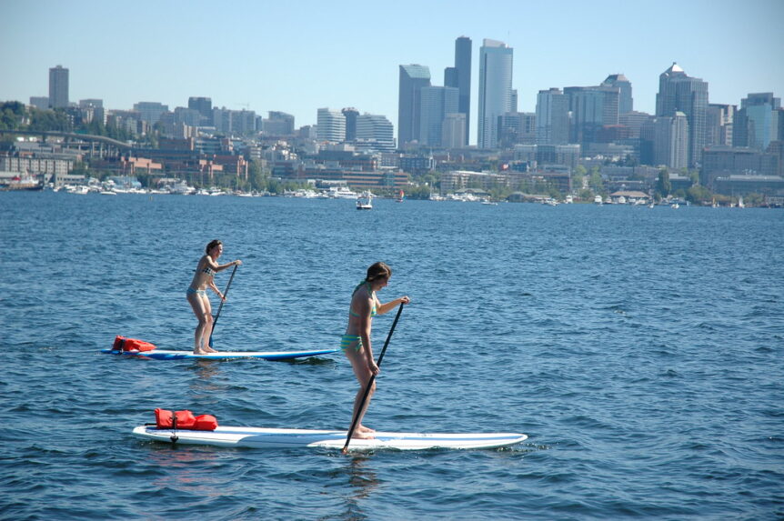 2 women on stand up paddleboards on a lake with a city in the backgroud